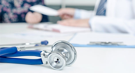 Stethoscope on a desk. Out of focus in the back you can see a doctor and patient